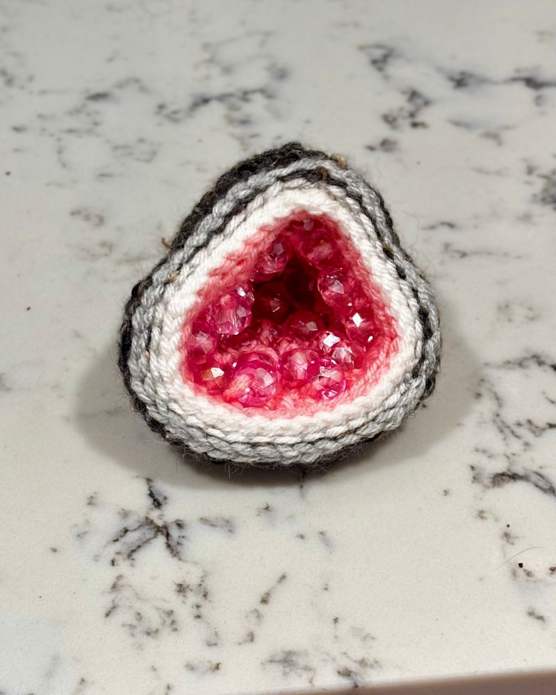 A soft crocheted geode made of yarn and pink beads on the inside to imitate crystals. It is sitting on a marble counter 