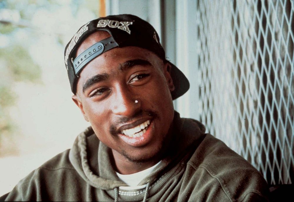An image of late hip-hop artist Tupac Shakur. He is smiling and looking off camera. He is wearing a backwards black baseball cap, a glittering nose stud, and a green hoodie while sitting in front of a chain link fence.