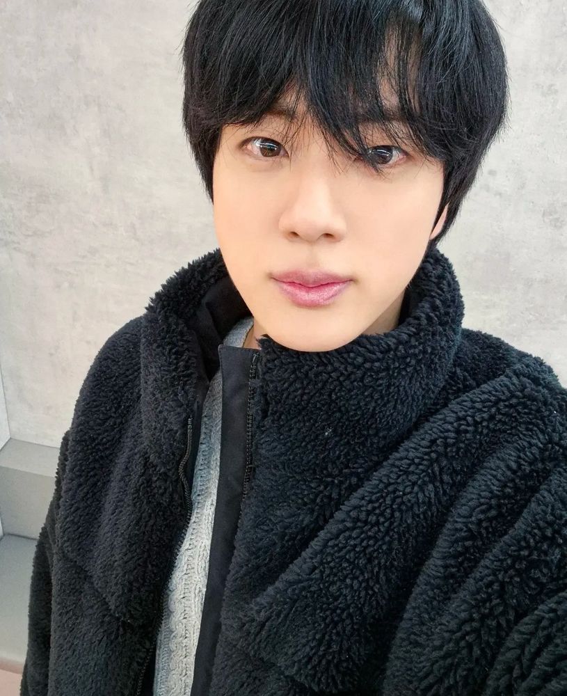 selfie of seokjin looking at the camera with noticeably longer hair since coming back and wearing a black fuzzy jacket