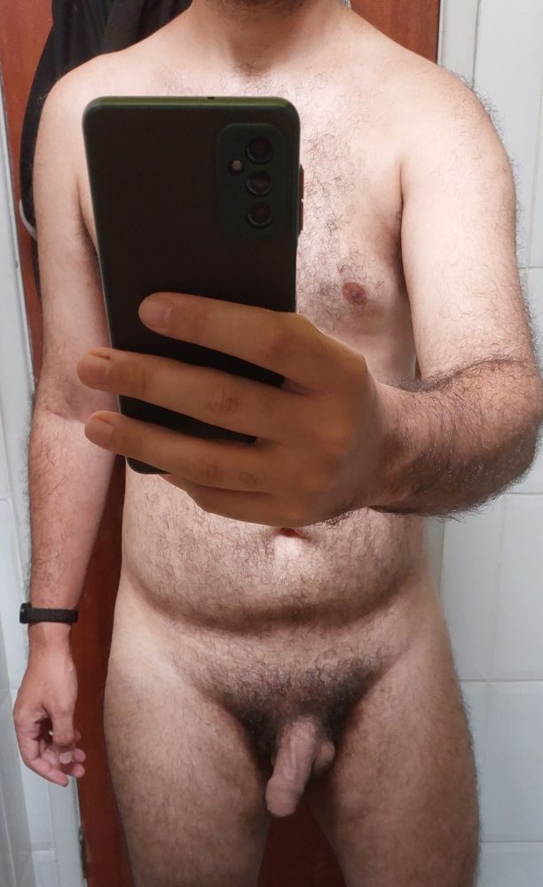 Um homem branco com pêlos no corpo segura o celular na frente de um espelho. Seu corpo é normal, com um penis quase flácido aparecendo. 