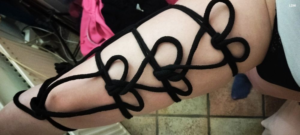 A photo of a leg with black rope and tied with hearts on the naked thigh.