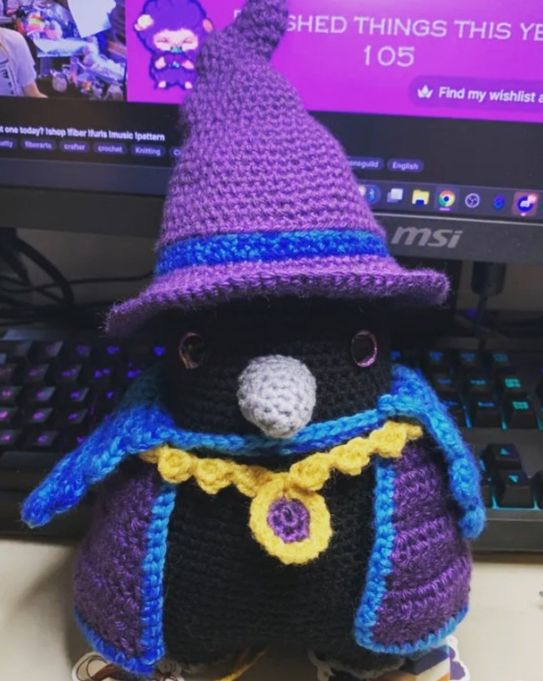 A black crochet raven with a gray beak, eyes with black pupils and sparkly purple irises, a purple irregularly-shaped witch hat with blue accents, a purple cape with blue accents, and a gold chain and amulet with a purple center (Pattern designer- CrochetedByBogusia on Etsy)