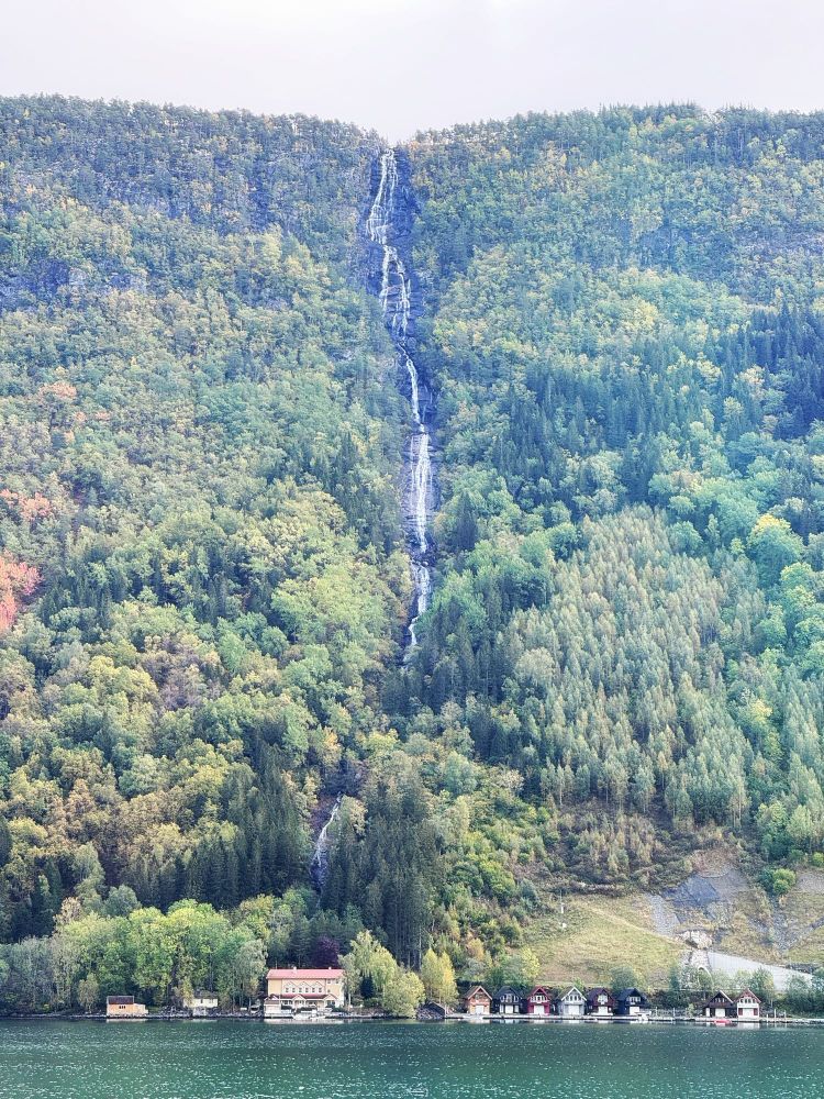 A tall, very thin waterfall down the side of a steep fjord above a house on the water. 