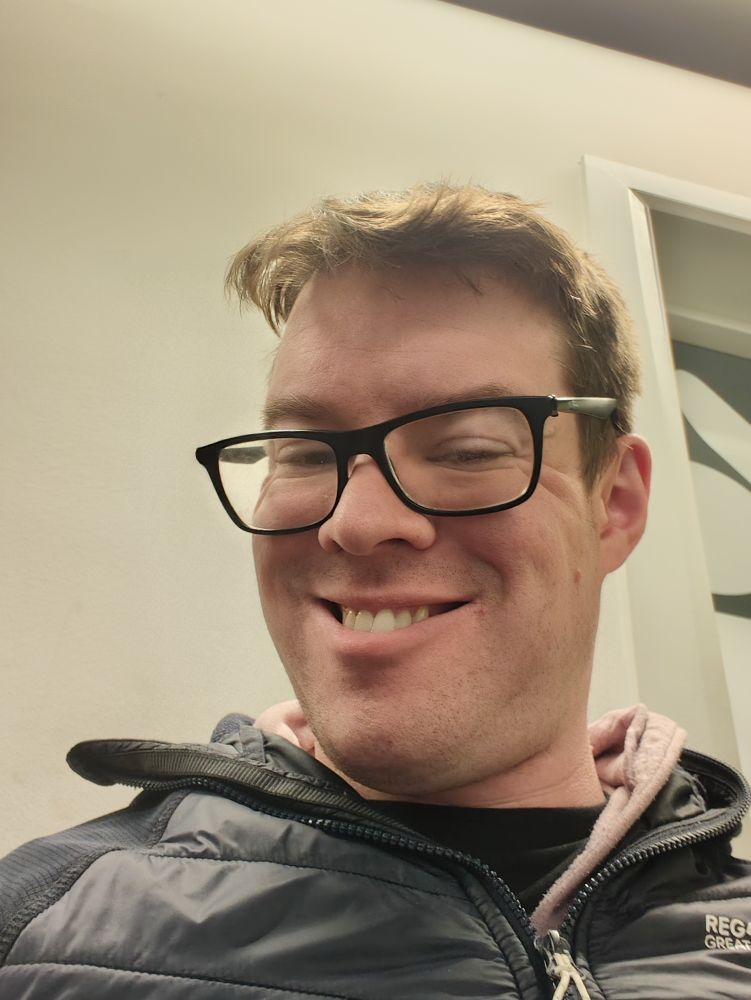 Person with brown hair and glasses, wearing a purple hoodie and a black puffer jacket. They're smiling.