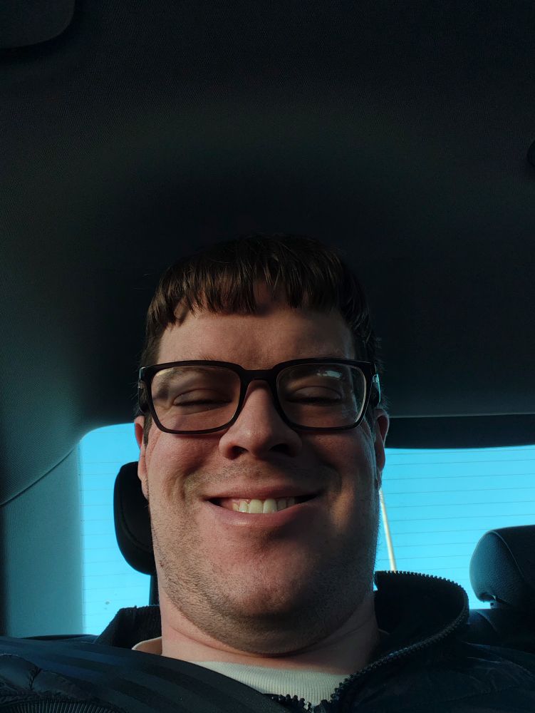 Person with brown hair and glasses wearing a blue puffer jacket, in the back of a car. They're smiling.