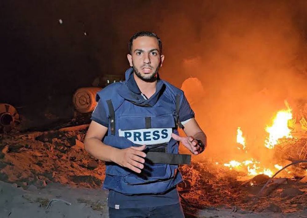 Anas Al-Sharif wearing a blue PRESS vest reporting in front of a bombed building on fire.