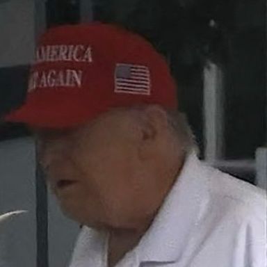 Trump in the shade at the WH wearing a white polo and red MAGA cap with his mouth agape.