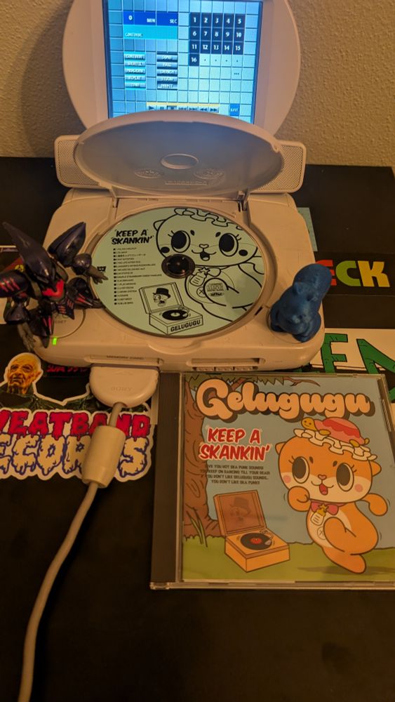 A PSOne with a flip up LCD screen with a CD inside. A small Qubeley and Squirrel are on the power and open buttons. The CD case for the CD under the PlayStation. The CD is Keep A Skankin' by Gelugugu. It has Chiitan in the cover dancing to what one can assume is the album playing on a record player. Chiitan is awesome. 