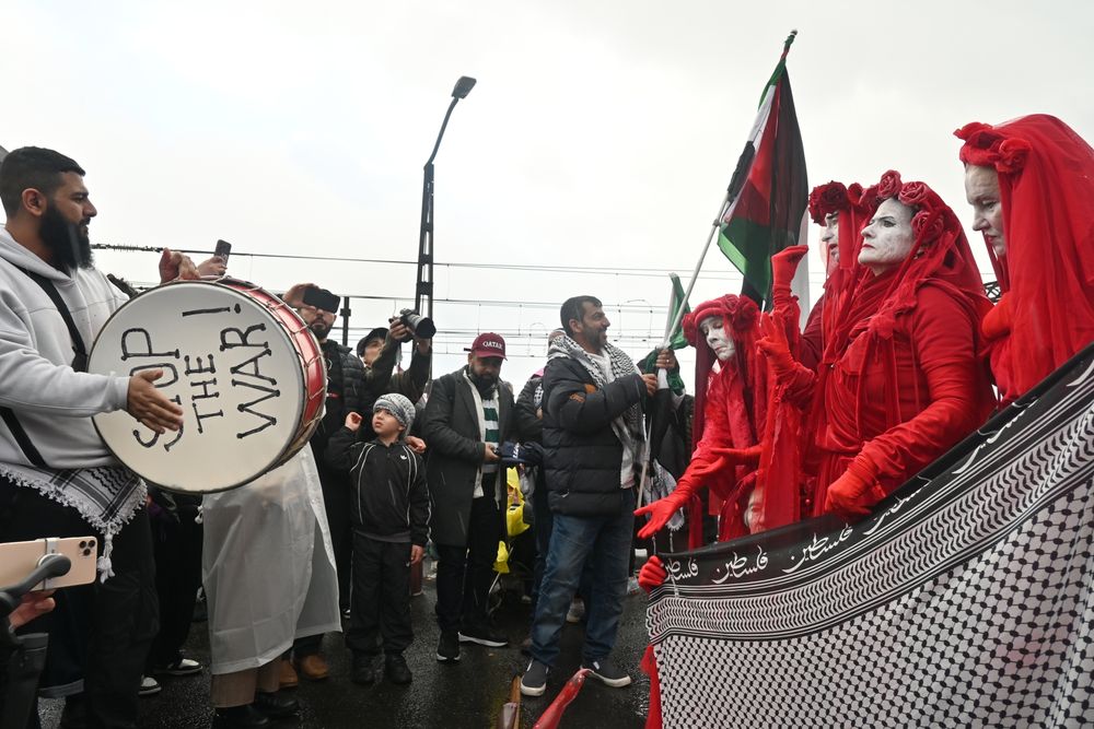A phot of protesters on the Sydney Harbour Bridge. A group of people dressed in red robes with white painted faces hold a black and white cloth with arabic text. A man plays a drum with his hands, the drum is strapped over his shoulders with the words "STOP THE WAR" on the drumskin. Young and old protesters stand around with Palestinian flags being waved.