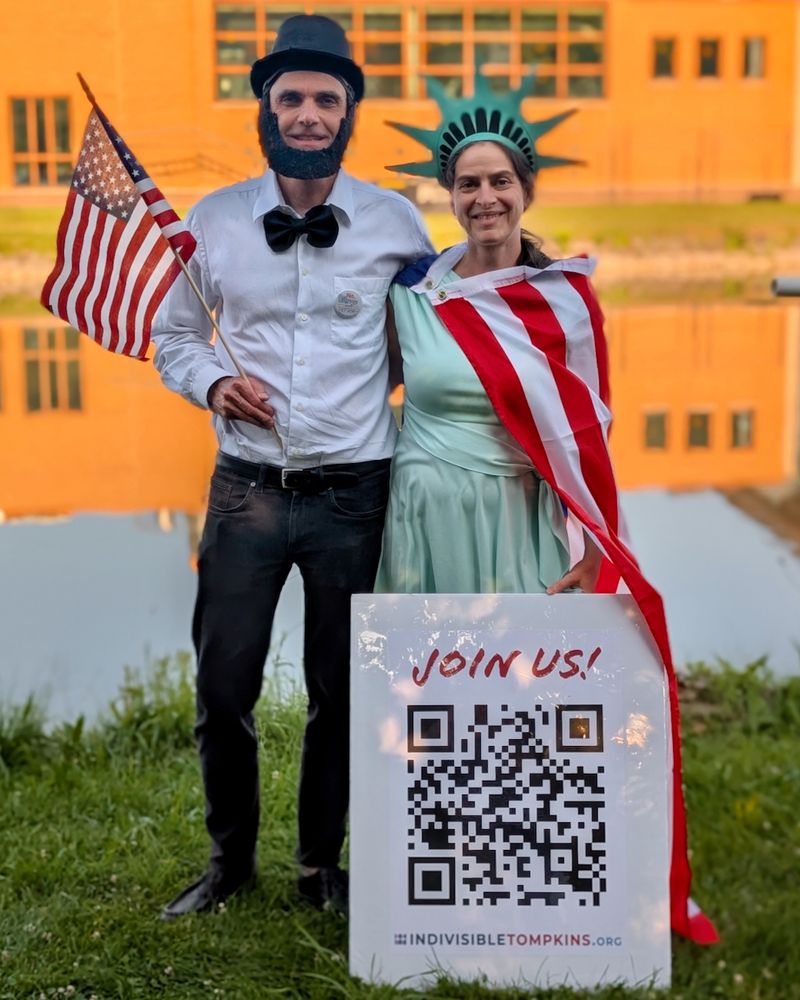 A couple dressed as Abe Lincoln and Lady Liberty, with a sign that reads "Join Us!" that has a QR code leading to indivisibletompkins.org