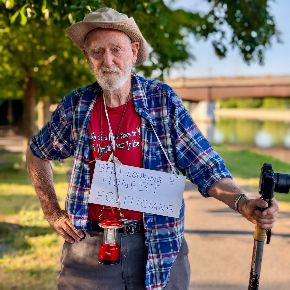 An older man in a blue flannel and tan hat with a sign reading "still looking 4 honest politicians."