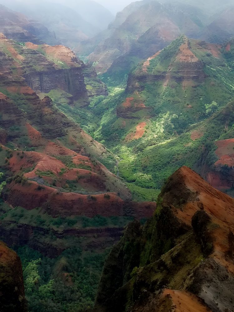 A breathtaking view of Waimea Canyon on Kauai, Hawaii, showing dramatic cliffs and ridges layered in red volcanic rock and lush green vegetation. Sunlight breaks through mist and clouds, illuminating the dense greenery at the canyon floor, while rugged slopes rise steeply around the valley, creating a vivid contrast of color and texture in the expansive landscape.