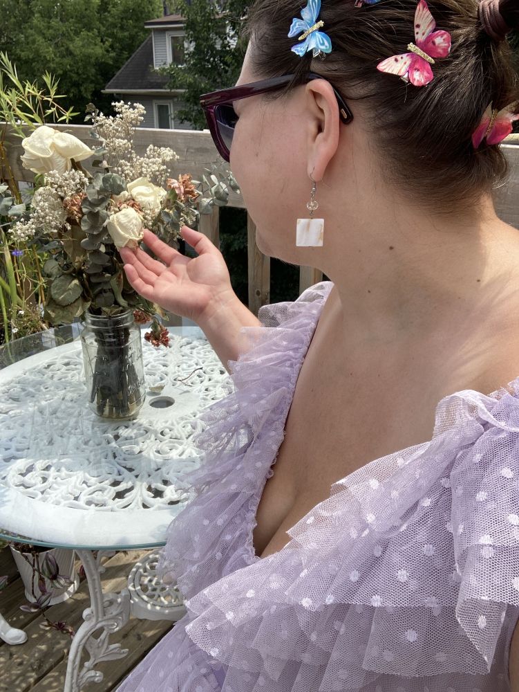 A brown haired woman with a collection of coloured butterfly clips in her hair wearing dark purple sunglasses and a fluffy light purple dress contemplates a bouquet of dried flowers.