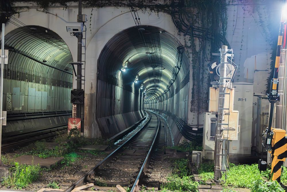 A picture of subway tunnels taken at night from ground level. Railings and lights curves inward and plants grow sparingly on the tracks.