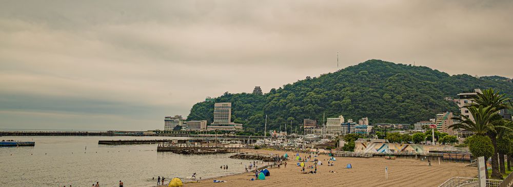 Photo of a beach with some people on it, some bathing. Slightly overcast, no blue sky visible. There is a mountain in the back with a temple visible in the center of the photo. Structures looking like hotels line the beach. Palms are visible to the right.