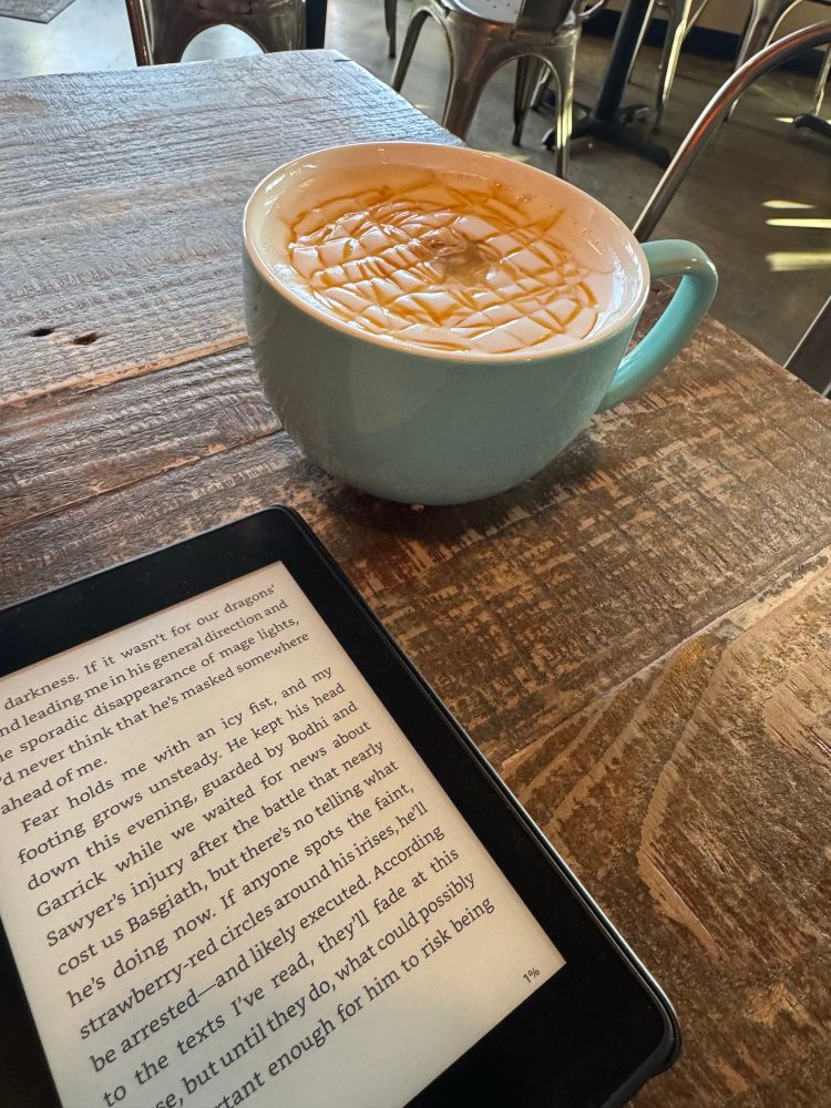 A kindle sits on a wooden table in front of a large teal mug filled with coffee. 