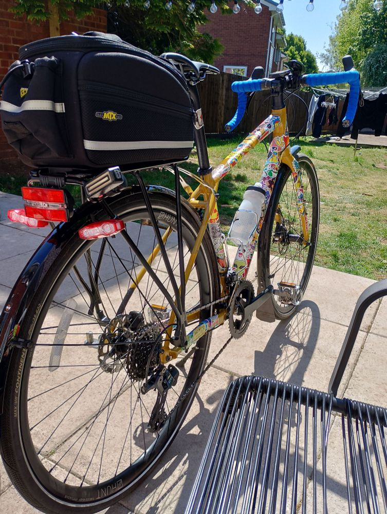 Picture of a yellow stickered gravel bike on touring tyres, with mud guards, luggage and a kick stand!
