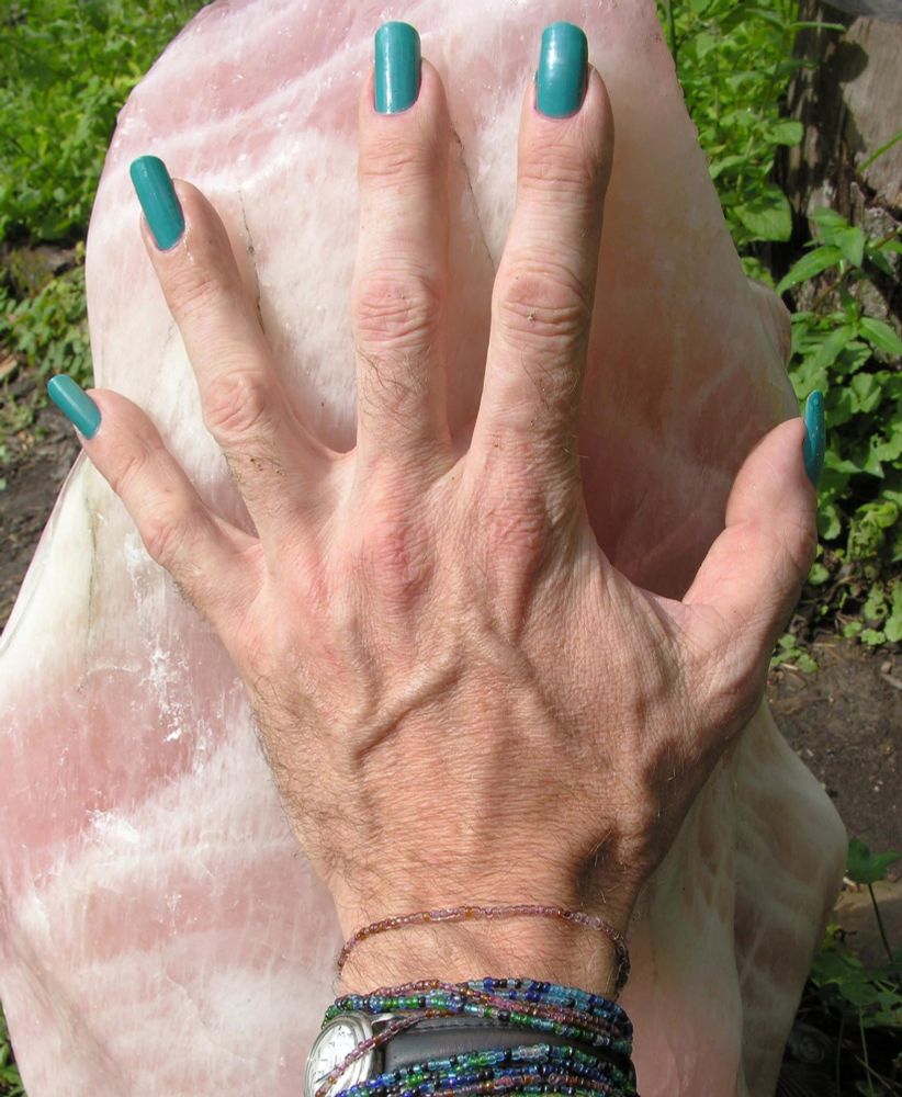 A large pale skinned hand with long turquoise painted nails against a large piece of rose quartz