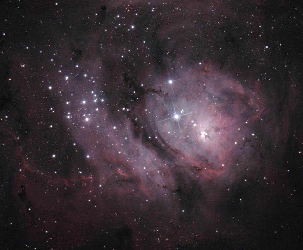 Image of the Lagoon Nebula, seen as glowing clouds of pink and bluish grey, with dark swirls and blobs and many stars of various sizes.