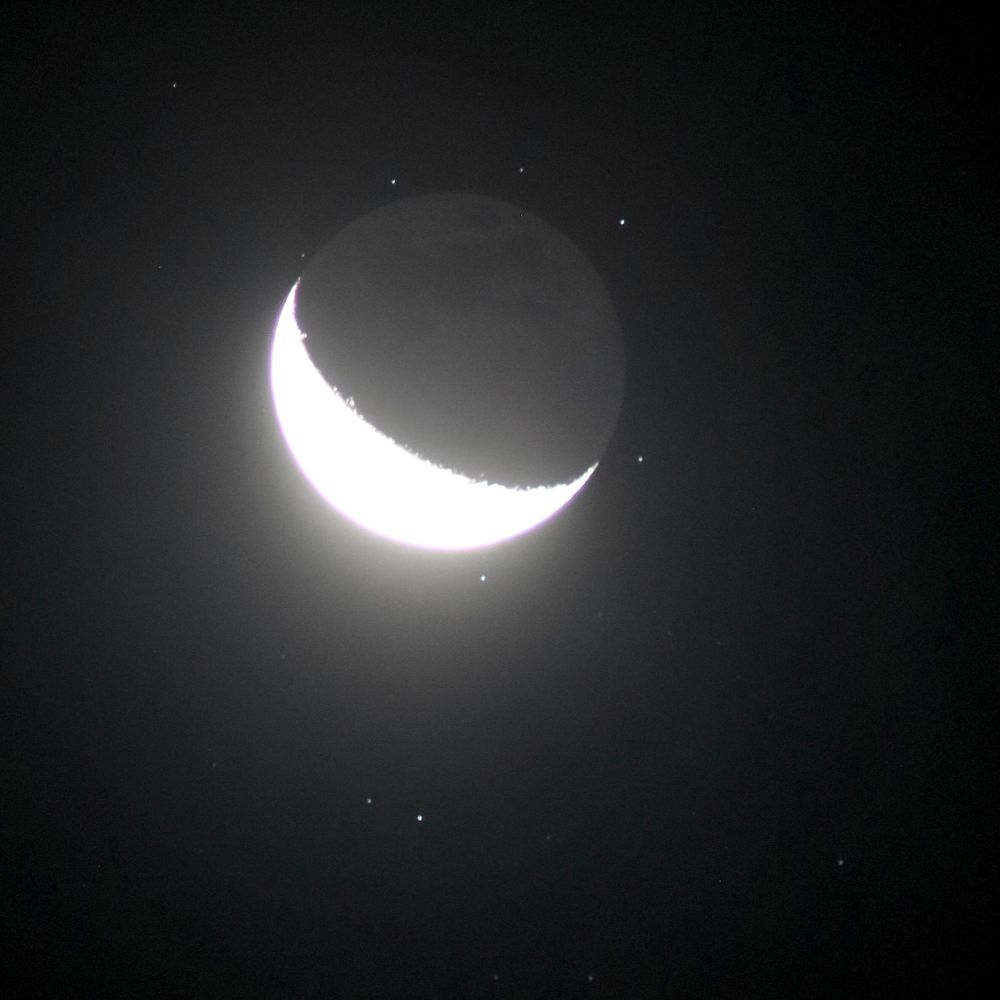 A very bright 23% crescent moon with faint earthshine on the dark side, surrounded by several small stars of the Pleiades cluster.