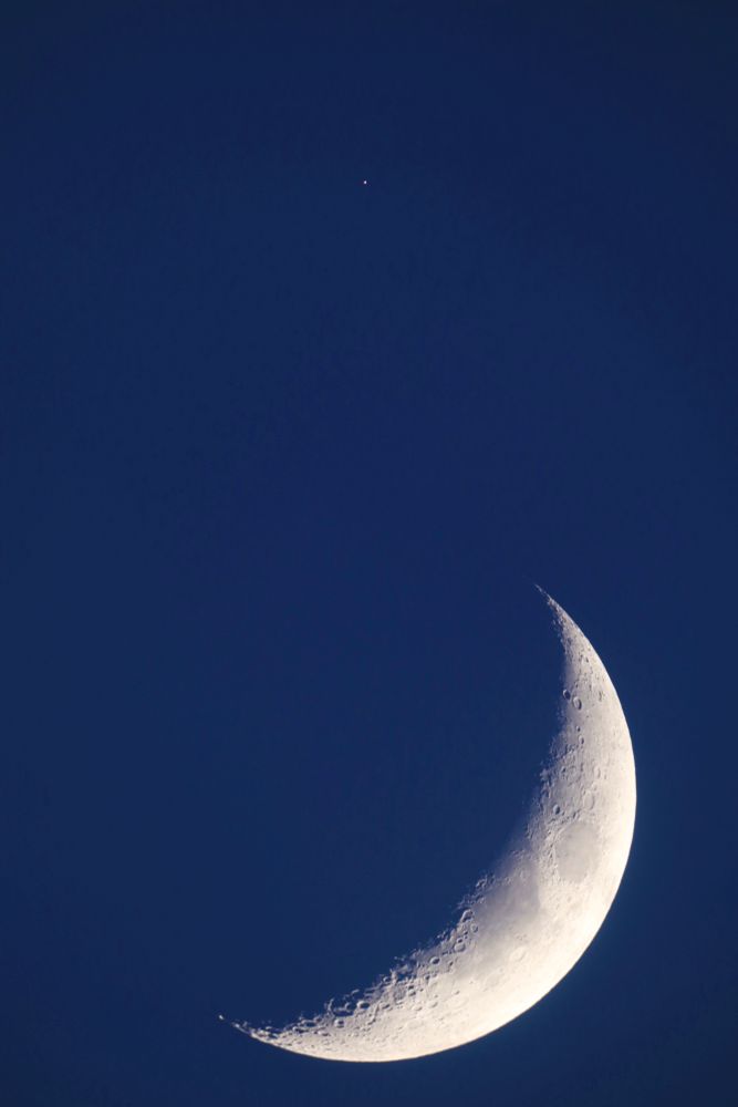 Crescent moon with some surface detail in dark blue sky, with a tiny point of light above, which is Mars