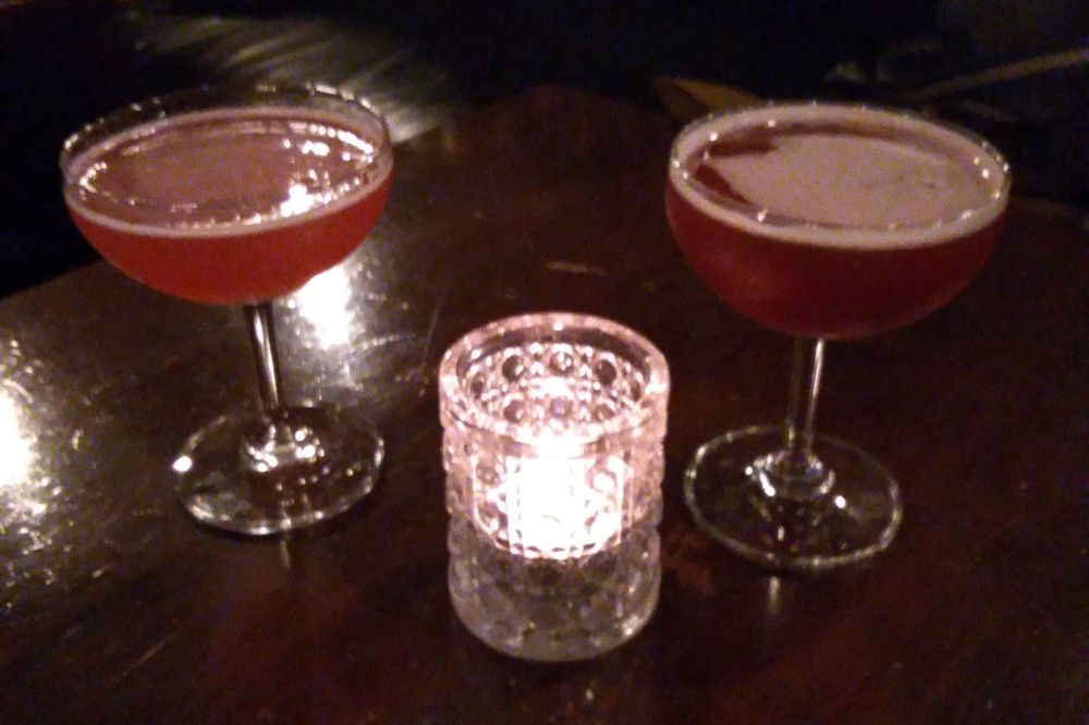 An indoor photo of two pink-red cocktails on a pub table with a lit candle in a glass.