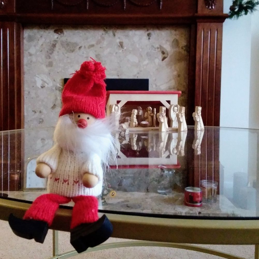 A toy santa sits on top of a glass coffee table. In the background is a wooden nativity and a fireplace.