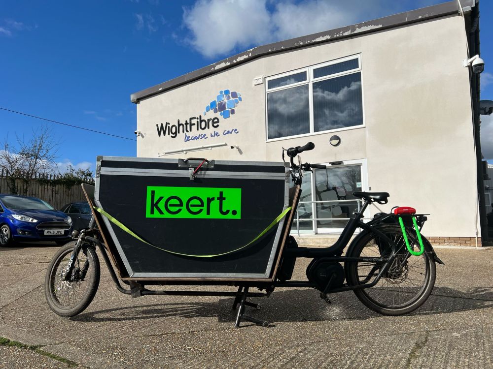A large black front loading cargo bike with bright green Keert logo on the box in front of a large white industrial building with a black and blue WightFibre logo, all under a gorgeous blue sky