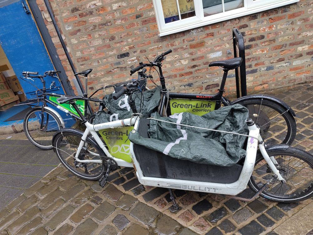 2 front loading Bullitt cargo bikes and one long tail Yuba cargo bike with Green-Link courier branding parked on a cobbled street in front of an old brick building with a blue door
