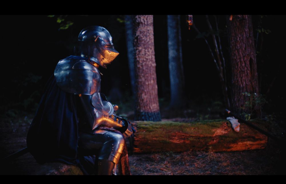 A screenshot from KNIGHTS & RELATIONSHIPS: a knight in armor sits by a fire at night.