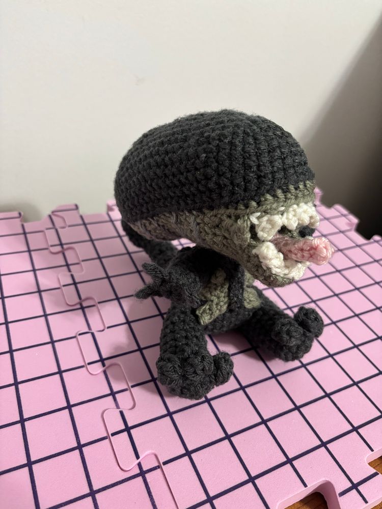 Crocheted xenomorph from Alien. The design is a cutesy version of the one from the movie. It is sitting and has its little arms out.