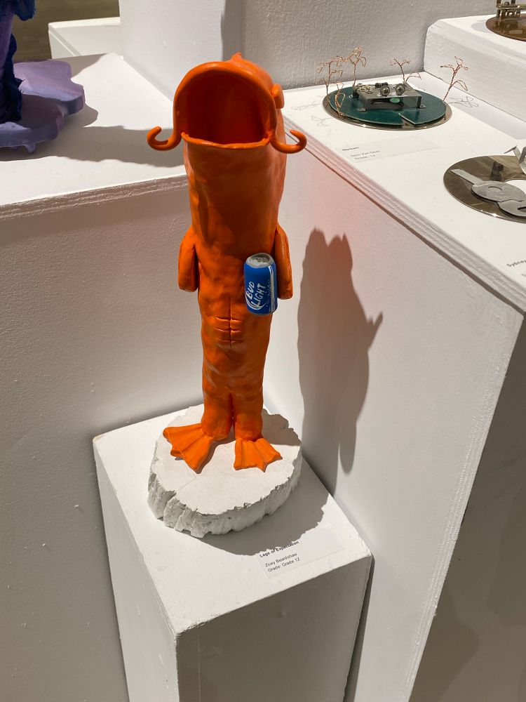 Photographed atop a white plinth is a ceramic student art sculpture of an anthropomorphic carp, holding a tiny can of Bud light. The carp has little ab muscles and a gaping mouth. 