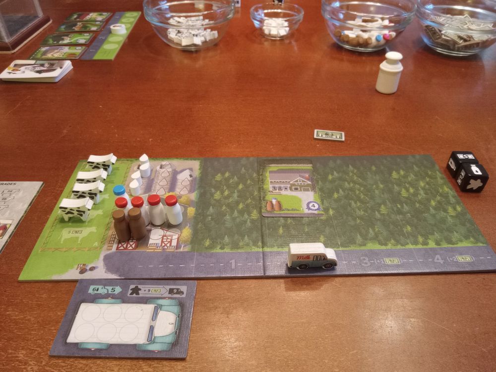 Milkman player board with milk truck, two chocolate milk bottles, four whole (red caps), one skim (blue), four raw milk tokens, and four cows. Various game pieces in background