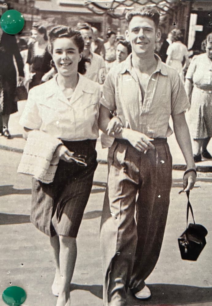 1950s. A young woman and man walk toward the camera with their arms linked. He is wearing double pleated wide leg trousers, a short sleeve pull over shirt and holding a camera case. She is wearing a pencil skirt and blouse. 