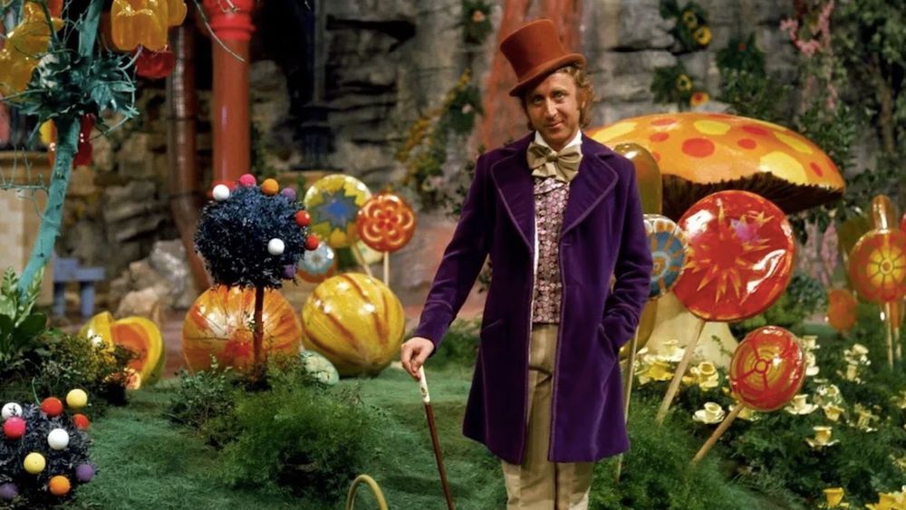 Still shot from the 1970s movie "Willy Wonka and the Chocolate Factory." Gene Wilder as Willy Wonka stands in a fantastical garden made entirely out of candy, wearing purple coat and orange top hat and leaning on a walking stick.
