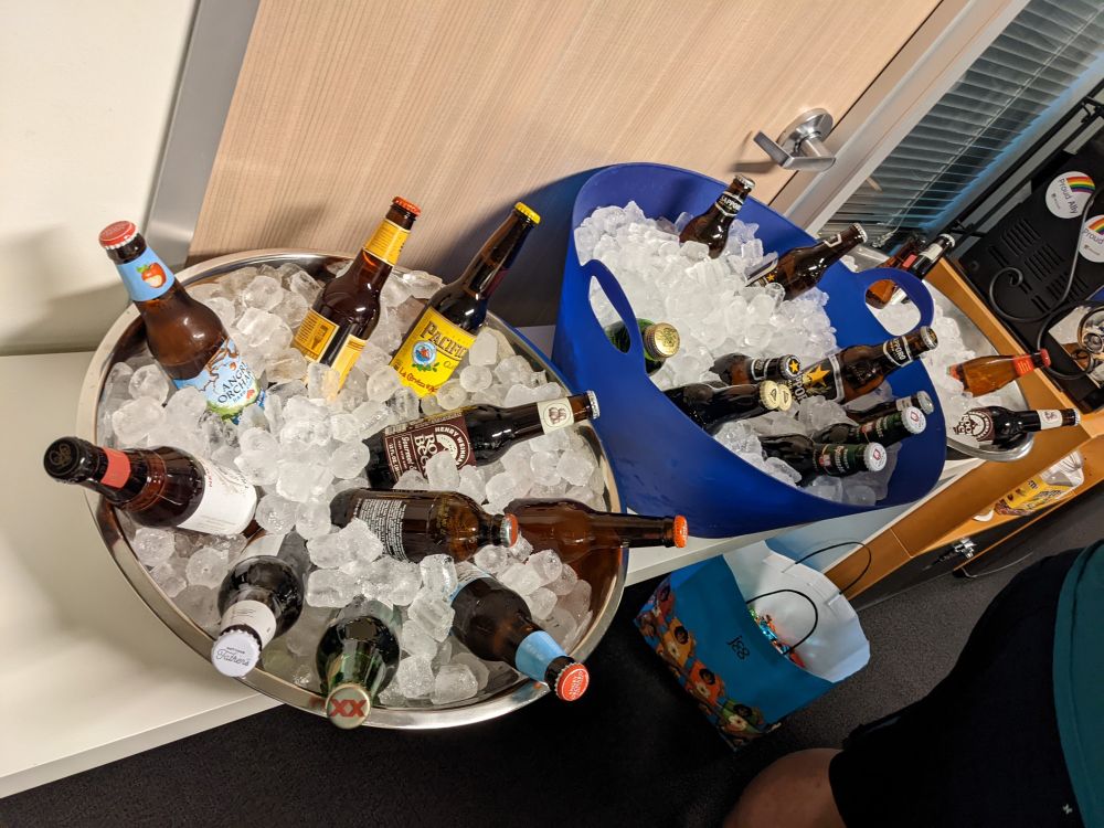 Three containers of ice with various beers and root beers in the ice. Two containers are metal and one is blue plastic. In the background is an office door and window, blocked by the table the beers are sitting on. Two "proud ally" magnets are visible on the side of the team microwave.