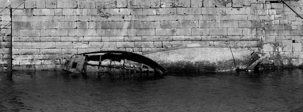 Black & white photo of 2 abandoned semi-submerged boat hulls in front of a stone wall