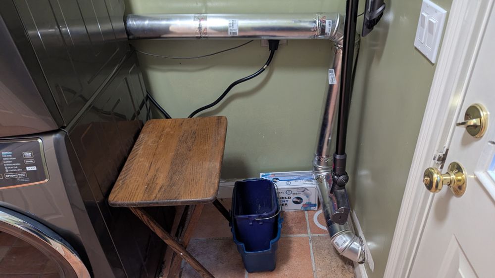 A rigid vent runs across a wall from a stacked washer/dryer, down the corner, across the floor, and out the exterior wall. A stick vacuum is mounted to the wall above the last segment of the run.