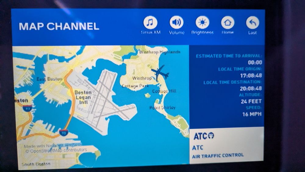 An in-flight entertainment screen showing a map of the area around Boston Logan Airport. There is an airplane icon on a peninsula across some water from the airport.
