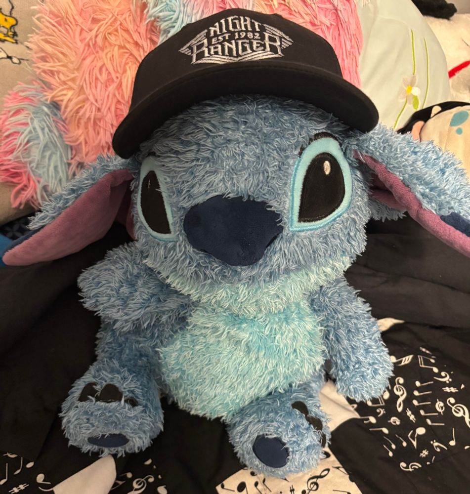 A blue Stitch plush sits on a black and white quilt with a musical note pattern on it. The Stitch plush is wearing a black baseball cap with a white logo in the middle of the front of the cap that reads Night Ranger. 