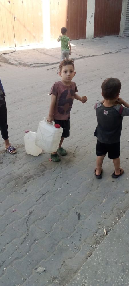 Rakan, a small brown haired boy, holding a big jug of water. His little brother, Mohammad, is next to him.