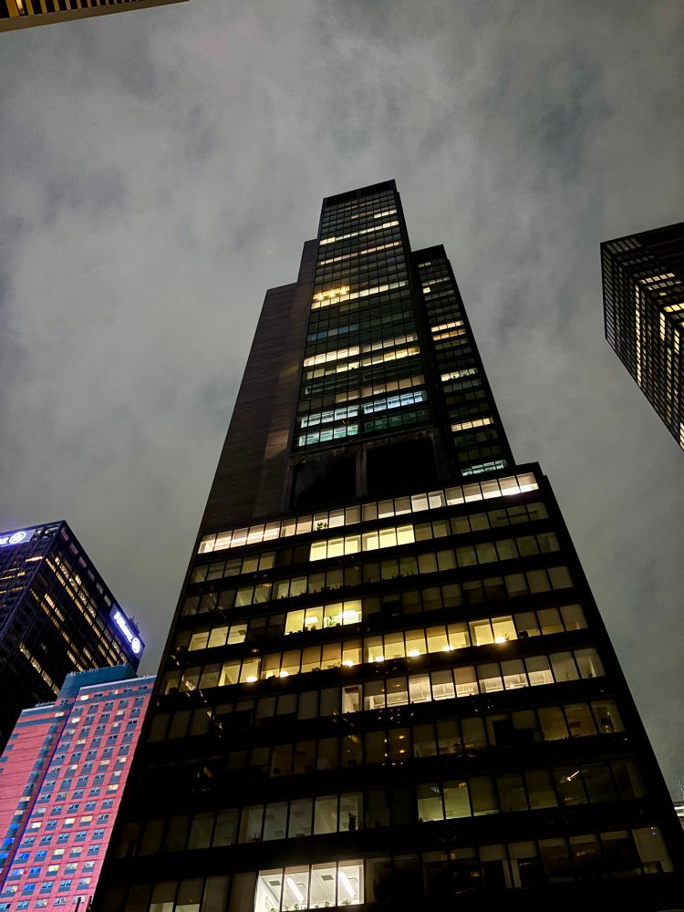 a skyscraper (?) with scattered lights light on each floor, a pink and blue building stands to the left, it is much smaller. the corner of another skyscraper leers in the background. the sky is many shades of grey, getting darker as it nears the corners of the frame