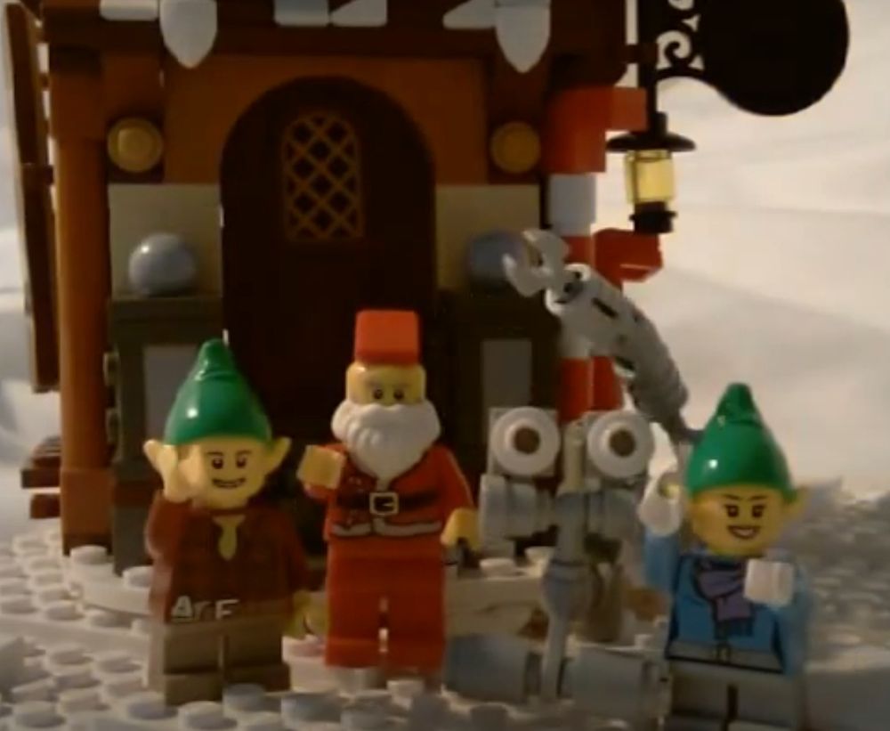 Lego Santa, elves and robot rover, standing in front of a Lego house, surrounded by snow.