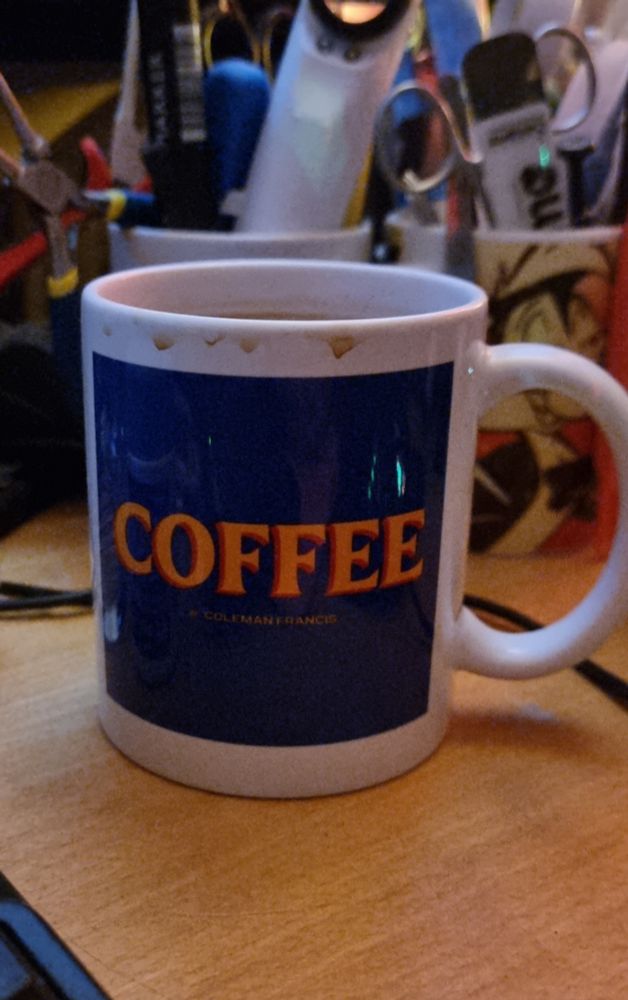 White coffee mug with a blue square and the word COFFEE written in a mustard colour. Top of the mug has many coffee runs from multiple uses without washing it up. Behind are slightly out of focus coffee mugs laden with tools - some forceps, a spudger, two types of needle nose pliers, a black marker pen, a magnifying glass with L E DEES. Too many electrostatical tweezles. A CARBON GRAPHITE prying tool, still in its packet because it's so fancy.