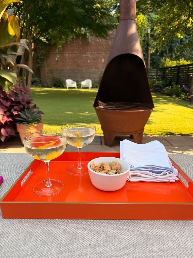 A photo of two martinis on an orange tray. In the background is a chiminea and a lush green yard. 