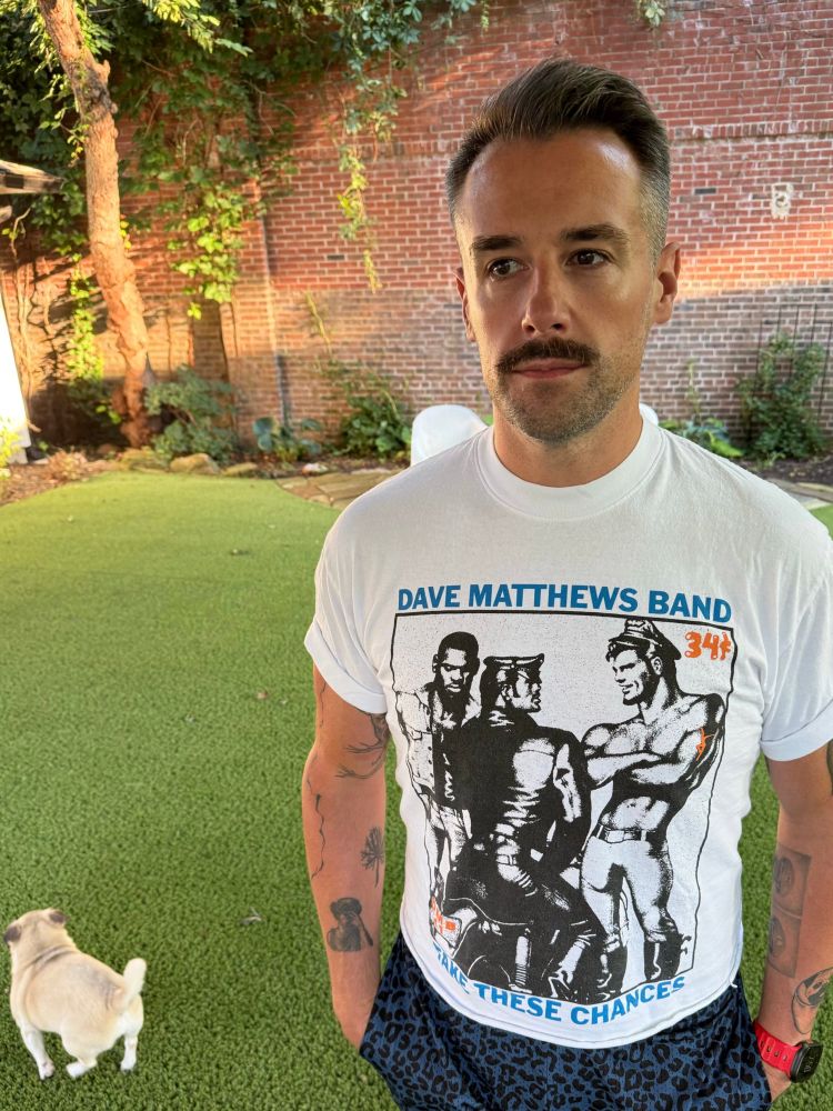 A man stares pensively into the distance. He is wearing a cropped t-shirt with Tom of Finland art on it, with a small pug in the background on green turf. 