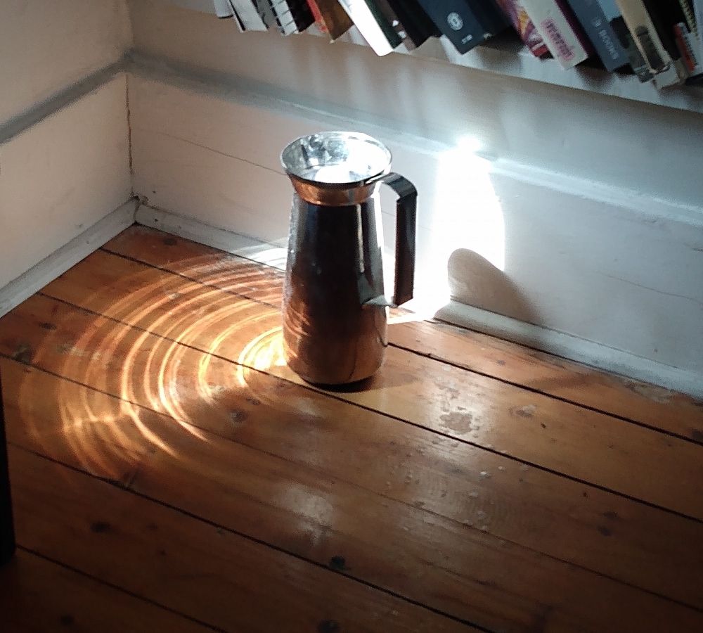 A metal jug on a wooden floor in the corner of a room under a low bookshelf. Sunlight is striking it making a water-like pattern of concentric rings on the floor around the vessel.