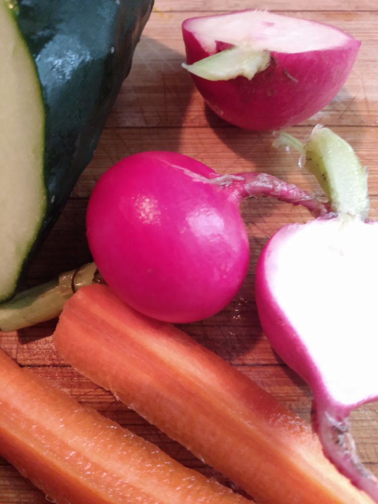 On a cutting board, fuschia radishes, one halved and white inside, orange carrot sticks,end of a green cucumber.