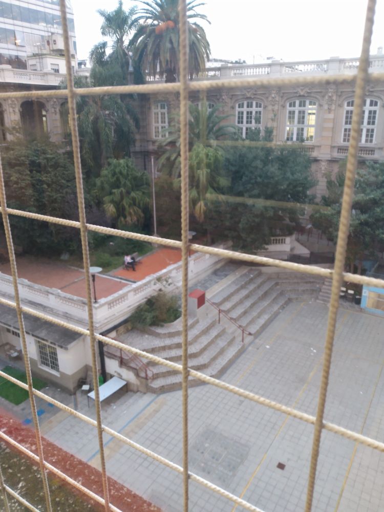 Through a latticed third floor window, a large gray stone courtyard with steps leading to a red tiled patio, large trees, another wing of the building across the yard.
