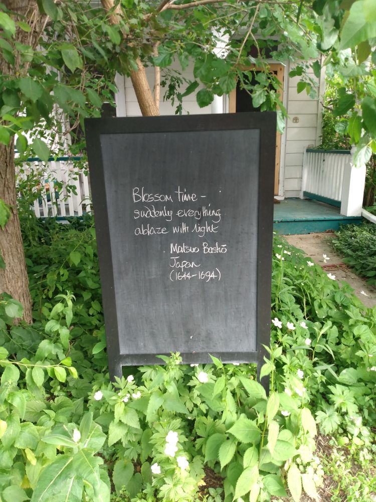A blackboard in the green and leafy front yard of a white-sidinged house says:

Blossom time -
suddenly everything 
ablaze with light.

Matsuo Bashō
Japan
1644-1694